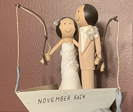 A handmade, wooden cake topper of a man and woman with dark hair, in a  boat that says NOVEMBER RAIN, each holding a reel with fish at the end of the line, and arms around each other.