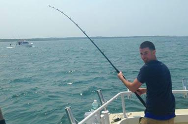 One of the guys standing at the edge of the boat reeling in a fish, looking back at the camera