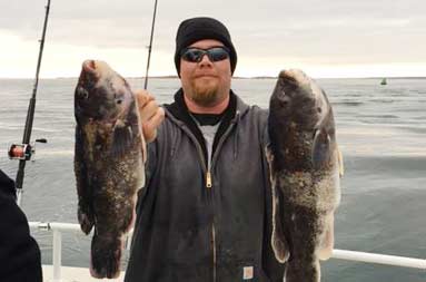 One of the guys in a coat and black wool hat holding up blackfish in each hand smiling into the camera