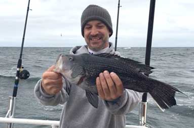 One of the guys in a sweatshirt and gray wool hat holding up the sea bass he caught