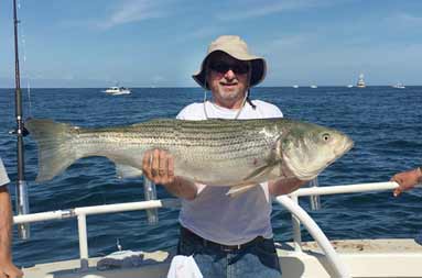 A man wearing a beige Helios sun hat and sunglasses holds up a large striped bass.