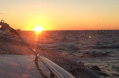 View through the boat's window with water droplets, looking out to the orange/yellow sunset ahead over the water.