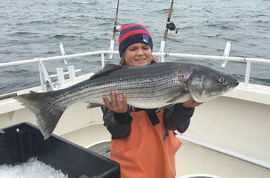 A young boy wearing a sweatshirt and hat proudly holds up a large striped bass.