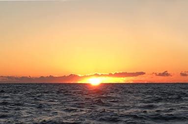 A sunset, with the vast ocean ahead and the sky with clouds in a yellowish-orange blue.