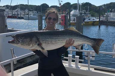 A woman smiles and proudly holds up the striped bass she caught.