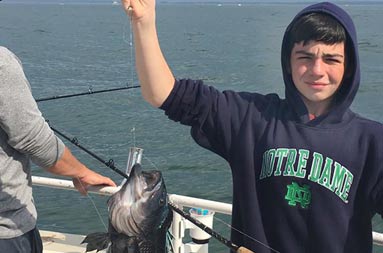 An adolescent boy wearing a dark blue hoodie sweatshirt holds up the sea bass he caught, still on the line.