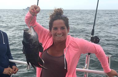 A woman in a pink hoodie sweatshirt proudly holds up the fish she caught.