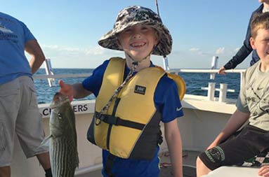 A young boy in a yellow life jacket proudly holds up the striped bass he caught.