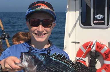 An adolescent boy smiles and holds up the sea bass he caught.
