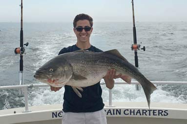 A young man, smiles as he holds up the striped bass he caught.