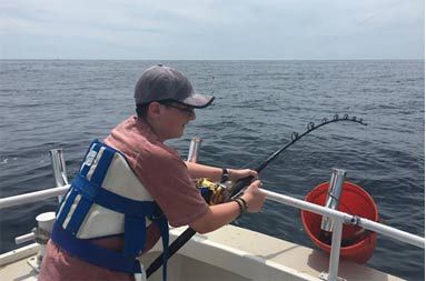 An adolescent boy holding onto the reel tight bent over the side of the boat as he fights a shark underwater on the other end.