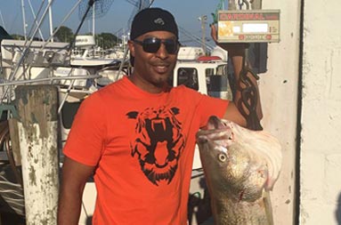 At the marina, a man in an orange shirt stands next to the scale where his fish hangs from. /><span class=
