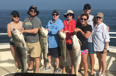 A group of 7 women and men hold up the 4 striped bass they caught.