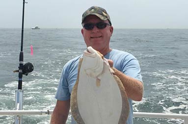 A man wearing sunglasses and baseball cap holds up his fluke.