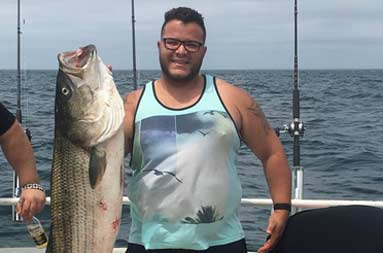 A man smiles and holds up the large striped bass he caught.