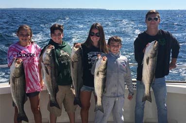 A man and 4 adolescent girls and boys smile as they each hold up the striped bass they caught. 