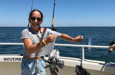 An adolescent girl smiles big as she holds up her reel with two sea bass on it.