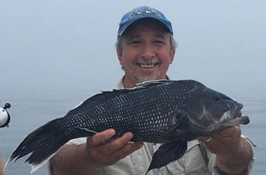 A man wearing a light blue baseball cap, smiles and proudly holds up the jumbo sea bass he caught for the camera.