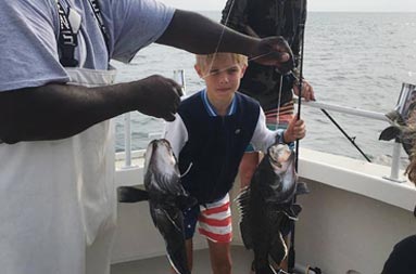 First mate, Gil, holds up the line with 2 sea bass attached as the young boy who caught them looks on
