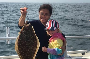A woman holds a fluke hanging on a hook with her armed around the young girl who caught it and is looking on.