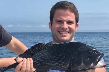 A man wearing a blue shirt holds out the jumbo sea bass he caught for the camera.