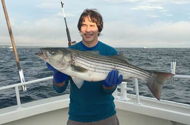 With the sunlight hitting his face and the clouds casting a shadow, a man holds up the large striped bass he caught.