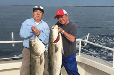Full length photo of father and son standing side by side smiling, with each holding up the striped bass they caught.