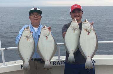 Father and son standing side by side smiling, each holding up the fluke they caught.