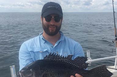 A man wearing a blue t-shirt and sunglasses holds up the sea bass he caught for the camera