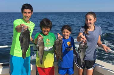 An adolescent boy and girl, along with a younger boy and girl each smile and hold out the sea bass they caught for the camera.