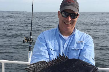 A man wearing sunglasses and gray baseball cap smiles big as he holds up his large sea bass.