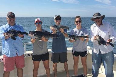 A father, along with his own father, and his set of triplet young adults, 2 women and 1 man, each hold up the sea bass they caught.