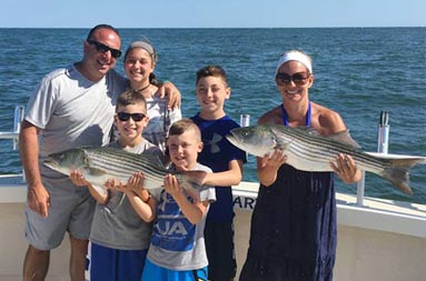 A woman holds up the striped bass she caught, with her husband, adolescent daughter and younger sons standing next to her. The 2 youngest sons are holding up another striped bass.