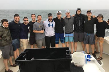 A group of 11 men gather around the boat and smile for a photo, on a rainy, overcast day.