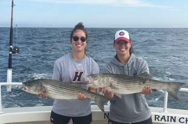 Twin sisters stand side by side smiling, each holding out the striped bass they caught.