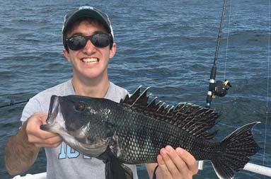 A young man smiles and holds up the sea bass he caught.