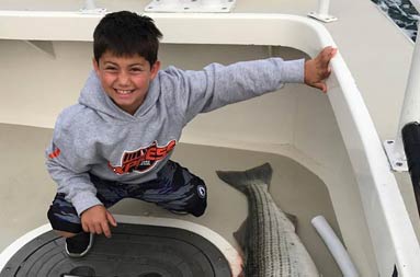A young boy smiles as he kneels down on the deck next to the striped bass he caught.
