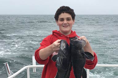 A teen boy wearing a red sweatshirt holds up 2 sea bass./><span class=