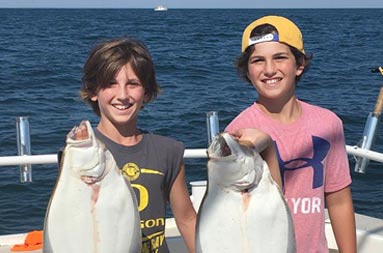 Two adolescent boys stand side by side and hold up the fluke they each caught.