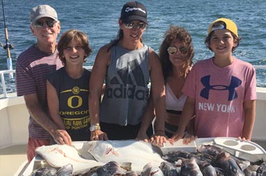 A family of 3 adults and 2 adolescent boys stand behind a spread of whole sea bass and fluke laid out on the fillet table.
