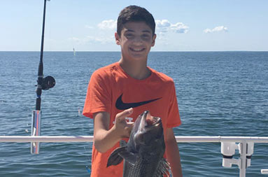 A teenage boy smiles big as he holds up the sea bass he caught.