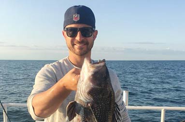 A man wearing sunglasses and a backwards dark blue baseball cap proudly holds up his sea bass for the camera.