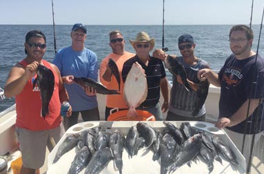 A group of 6 men ranging in age, hold up their catch as they stand around the fillet table where whole sea bass are spread out.