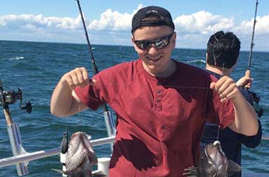 A man wearing a maroon t-shirt, black baseball cap and sunglasses holds up 2 sea bass.