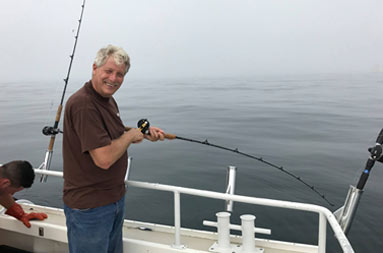 With a foggy, overcast sky in the background, a man faces the camera as smiles as he waits for a bite on his line.