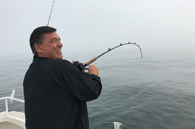 With a foggy, overcast sky in the background, a man looks back at the camera and smiles as he waits for a bite on his line.