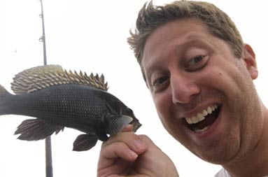 A closeup of a man smiling, holding a very small sea bass next to his face.