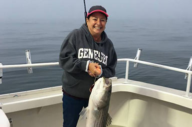 A woman holds up a striped bass by the hook, and blood covers some of the deck directly below the fish.