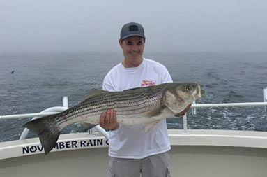 A man smiles and holds up a striped bass with both hands.