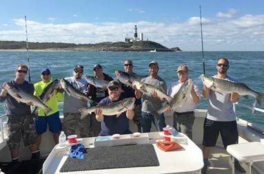 With the Montauk lighthouse, along with a blue sky and clouds in the background, 9 men smile and each hold up the striped bass they caught .